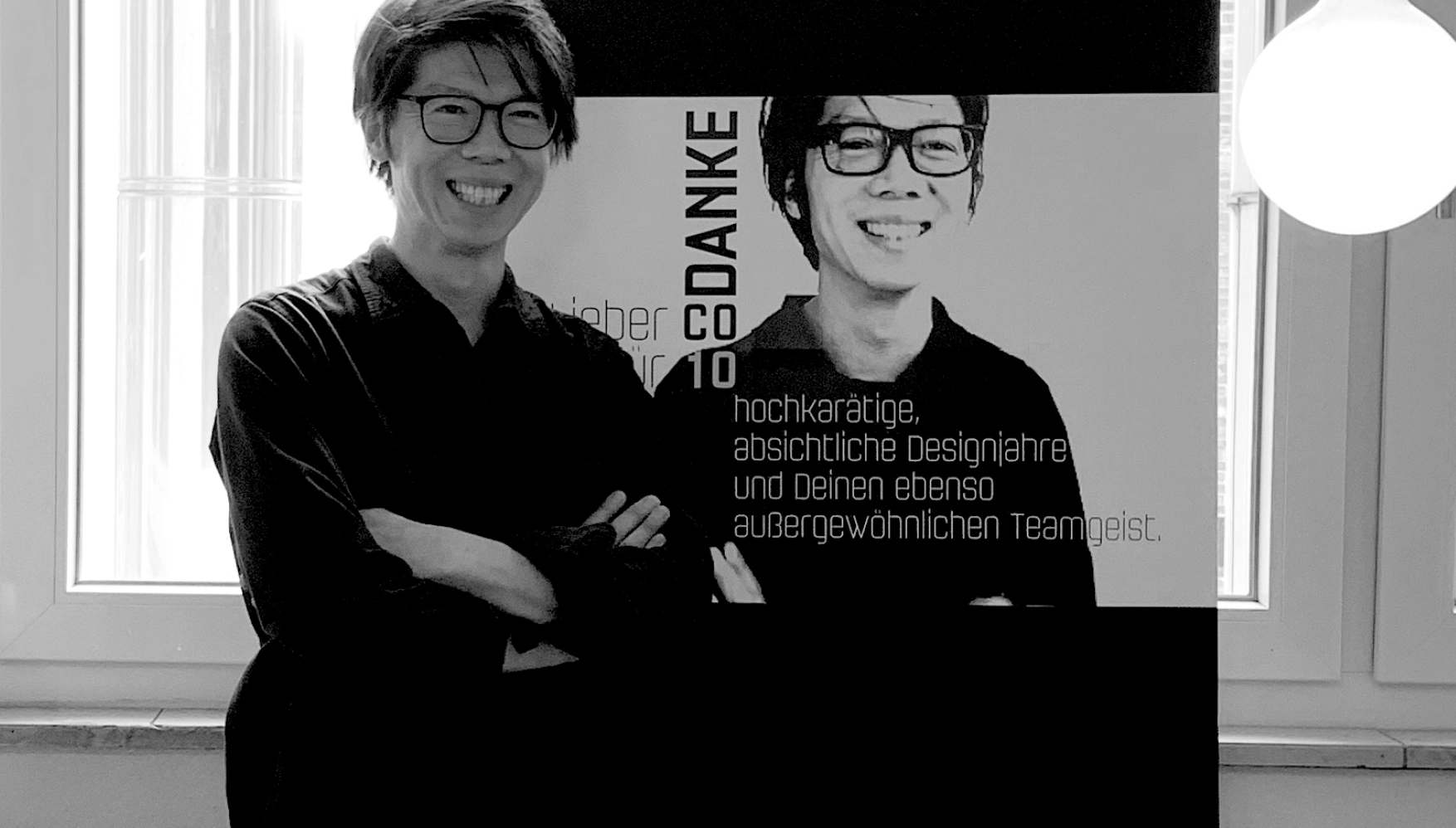 Smiling person with glasses standing with arms crossed in front of a poster showing another smiling person and German text thanking for 10 years of design and team spirit.