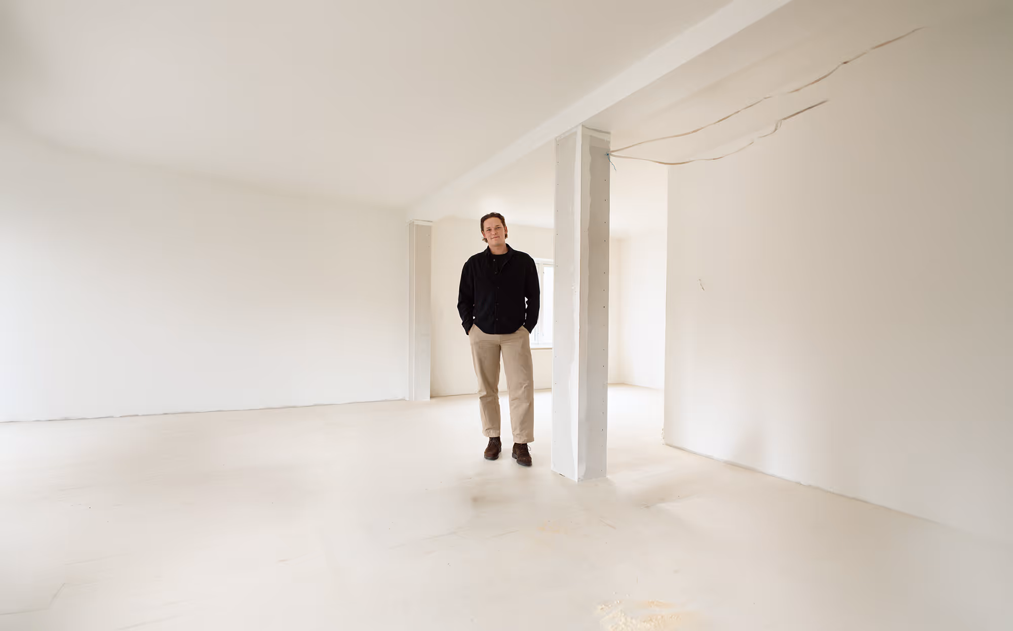 Mand iført sort jakke og beige bukser står med hænderne i lommerne i et tomt hvidt rum med søjler.