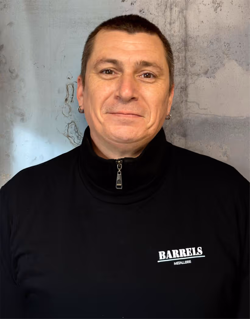 Smiling man with short dark hair wearing a black zip-up sweater with 'Barrels Metallerie' logo, standing against a textured gray wall.