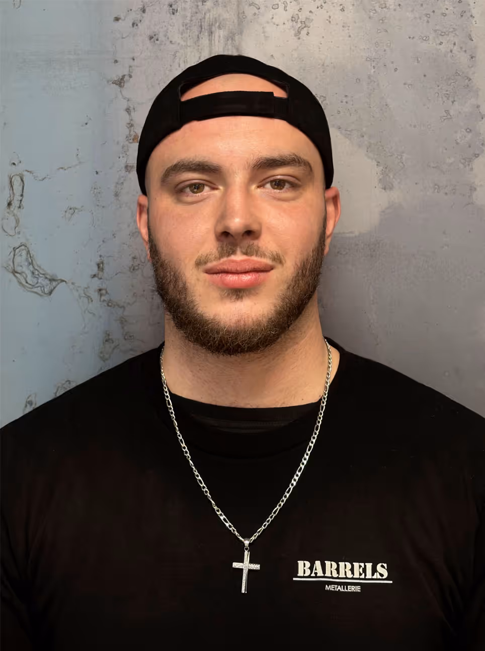 Young man with beard wearing a backward black cap, black shirt with 'BARRELS METALLERIE' logo, and a silver cross necklace.
