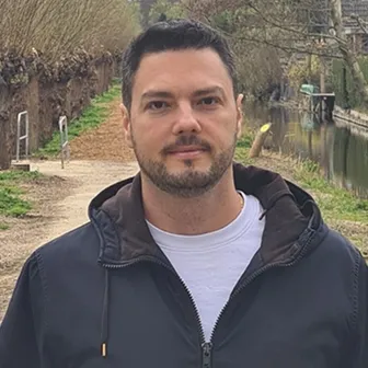 Man with short dark hair and beard wearing a dark jacket over a white shirt standing on a riverside path with trees in the background.