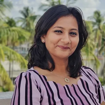 Smiling woman with shoulder-length black hair wearing a purple and black striped top, with palm trees blurred in the background.