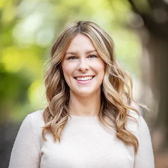 Smiling woman with long wavy blonde hair wearing a light-colored sweater outdoors with blurred green and brown background.