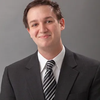 Smiling man in a dark suit and striped tie against a gray background.