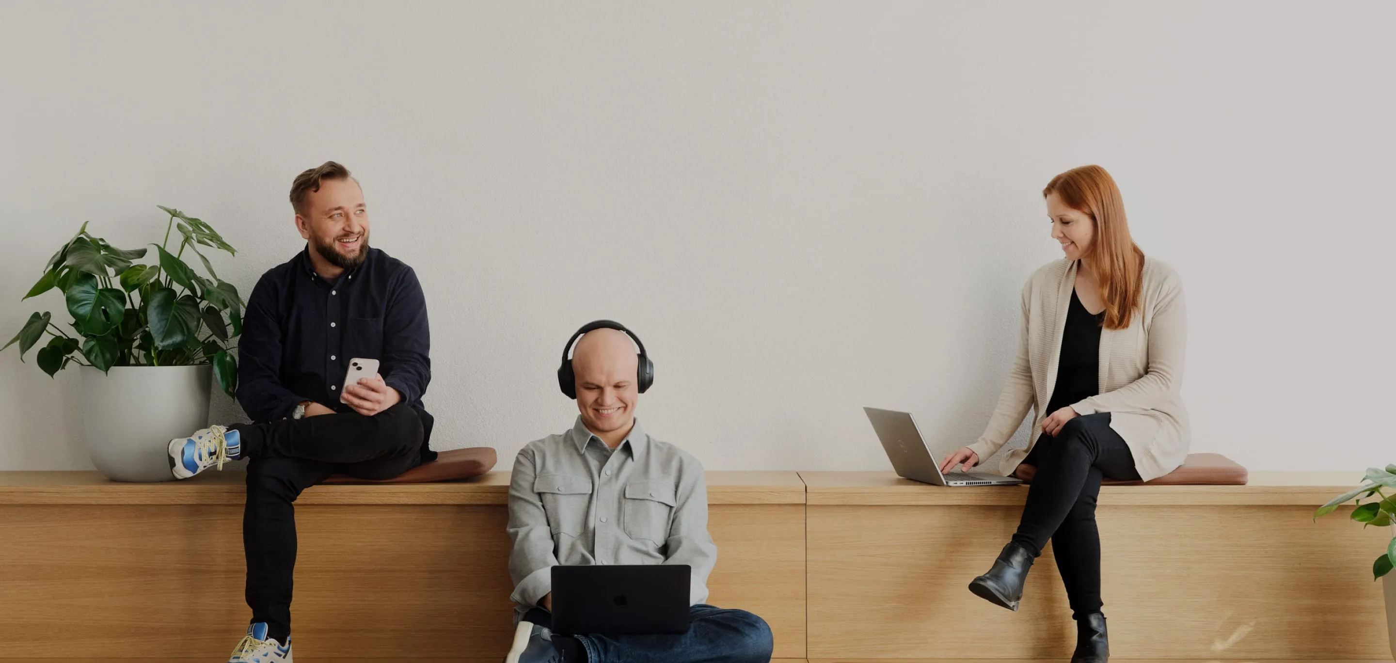 Three people in casual attire working with laptops and a smartphone while sitting on or near a wooden bench with potted plants on each side.