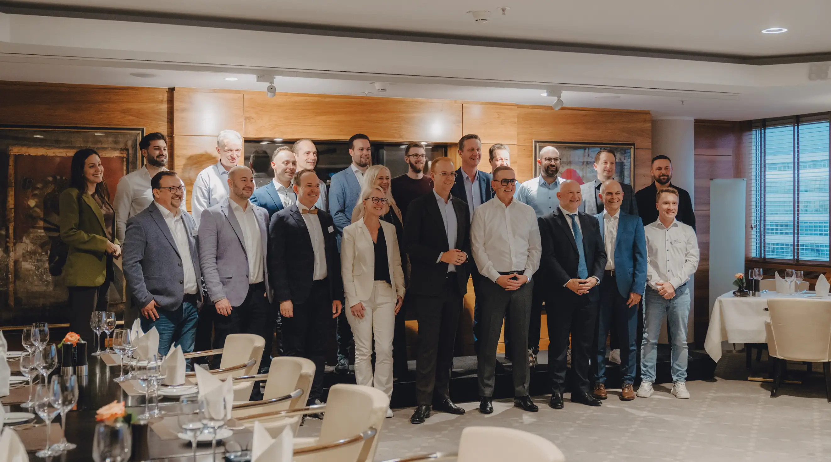 Gruppenfoto von 20 Personen in Geschäftskleidung in einem hellen Konferenzraum mit verglasten Fenstern und gedeckten Tischen.