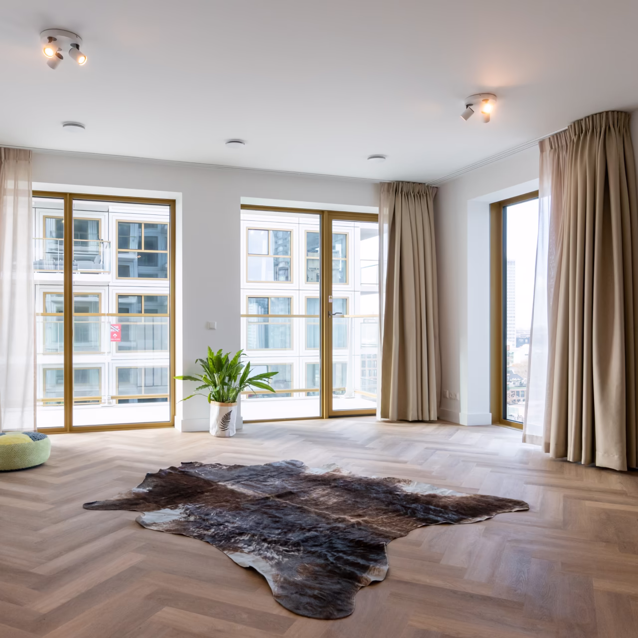 Lichte, lege kamer met houten visgraatvloer, een dierenhuidtapijt, een groene plant en grote ramen met beige gordijnen.