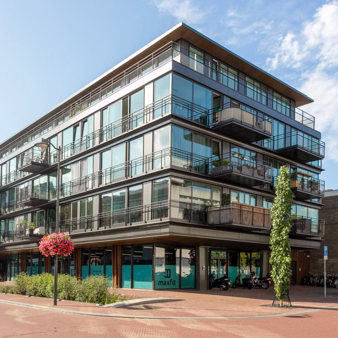 Modern hoekappartementengebouw met grote glazen ramen en balkons, gelegen aan een stille straat met bloeiende boom en fietspaden.