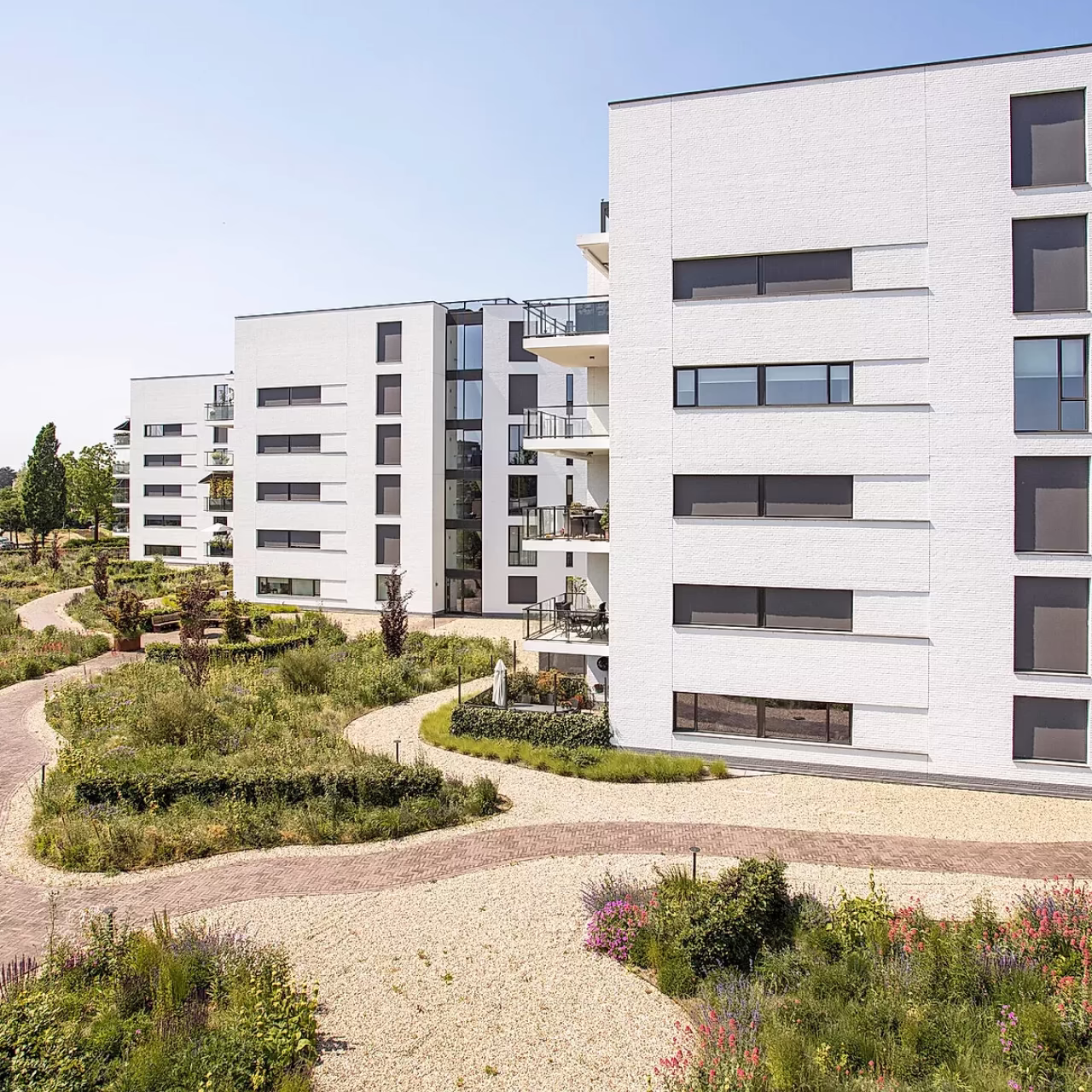 Moderne witte appartementencomplexen met meerdere verdiepingen en smalle ramen, omgeven door een tuin met wandelpaden en groen.