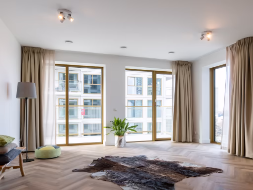 Lichte, ruime woonkamer met grote ramen, houten vloer, groene plant en beige gordijnen.