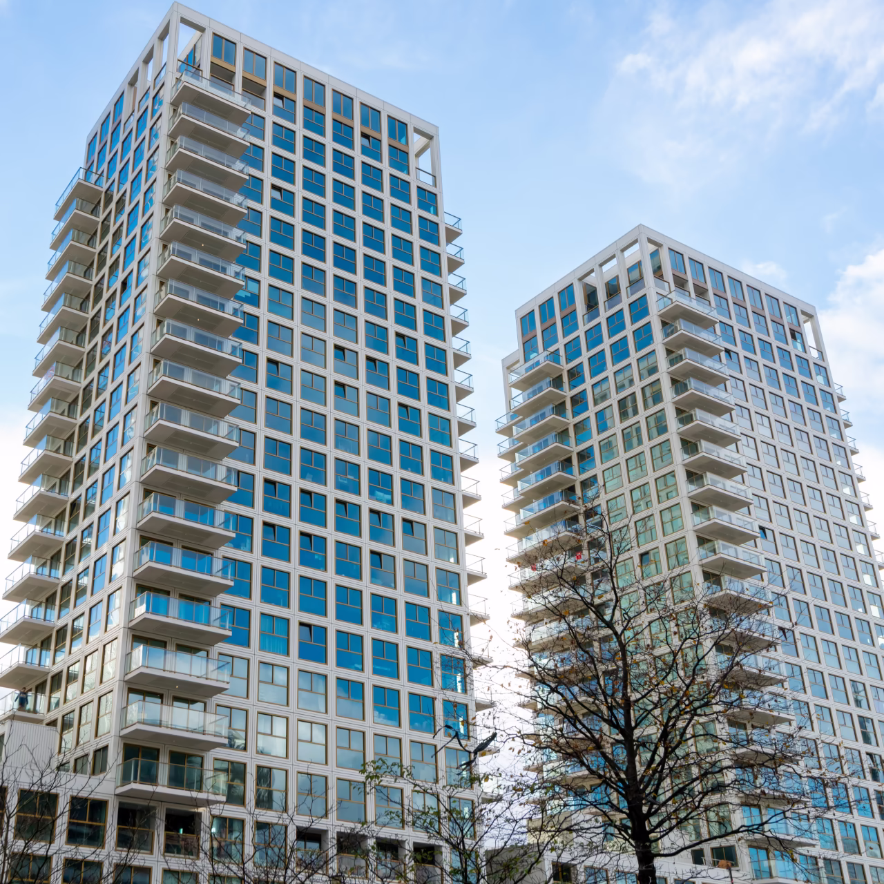 Twee moderne hoge woongebouwen met glazen balkons tegen een blauwe lucht met enkele wolken.