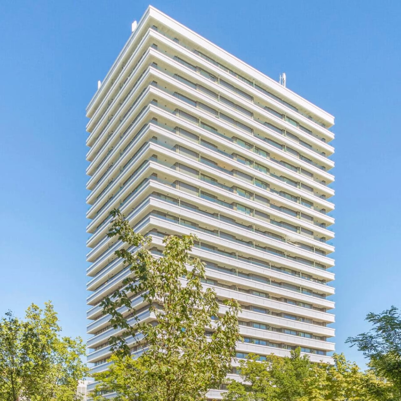 Hoog appartementengebouw met balkons en bomen op de voorgrond onder een heldere blauwe lucht.