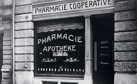 Schwarz-weiß-Foto einer Apothekenschaufensters mit der Aufschrift ‚Pharmacie Apotheke‘ und ‚Pharmacie Cooperative‘ darüber.
