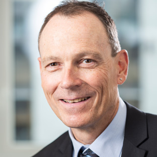 Mittelalterlicher Mann mit dunklem Anzug und Krawatte lächelt vor einem unscharfen Hintergrund.