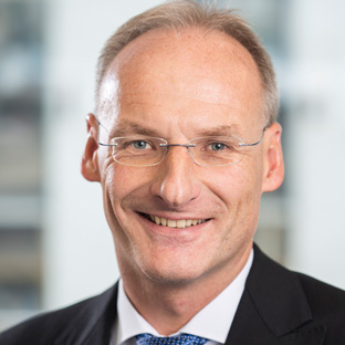 Porträt eines lächelnden Mannes mittleren Alters mit Brille, kurz geschnittenem Haar und Anzug vor unscharfem Hintergrund.