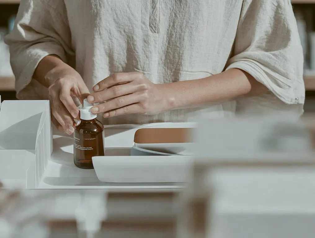 Person dreht an einem braunen Glasfläschchen mit weissem Deckel auf einem Tisch mit weissem Hintergrund.