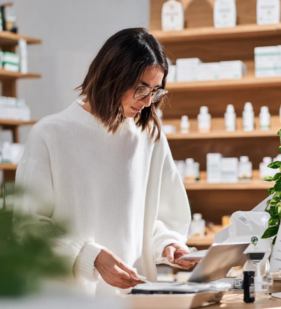 Frau in weißem Pullover prüft Produkte in einem Geschäft mit Regalen voller Flaschen und Kartons im Hintergrund.