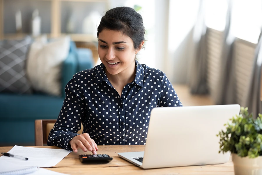 Woman sitting at a table using a calculator with a laptop and paperwork in front of her, smiling.