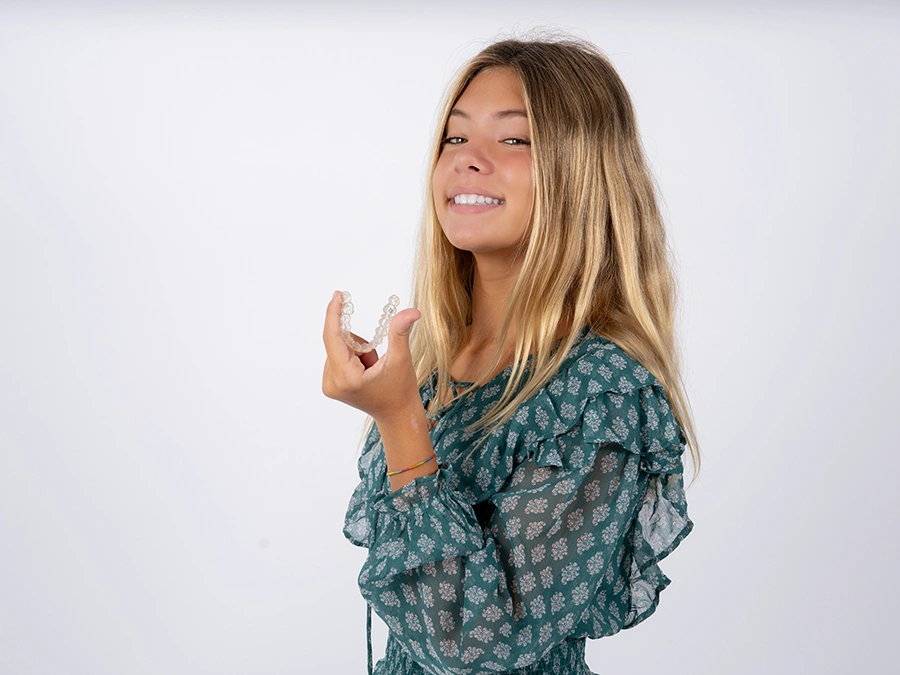 Smiling teenage girl with long blonde hair holding a clear dental aligner against a plain white background.