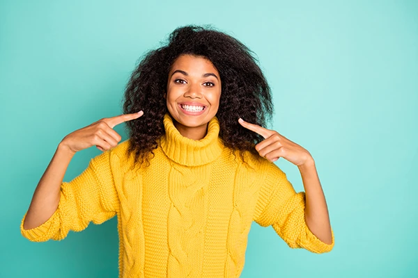 Smiling woman in a yellow sweater pointing at her bright white teeth against a turquoise background.