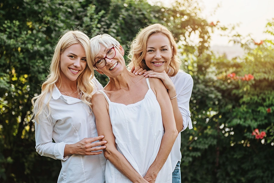 Three smiling women from different generations standing closely together outdoors with greenery in the background.