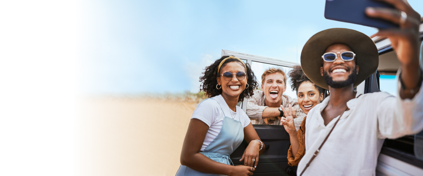 Four friends smiling and taking a selfie outdoors near a vehicle on a sunny day.