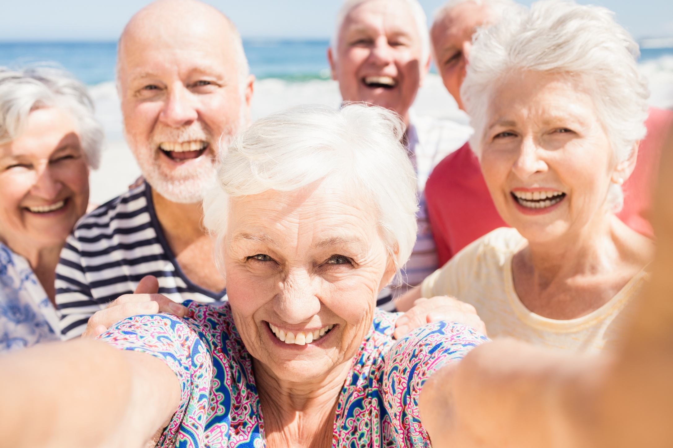 Group of six smiling elderly people taking a selfie at a sunny beach.
