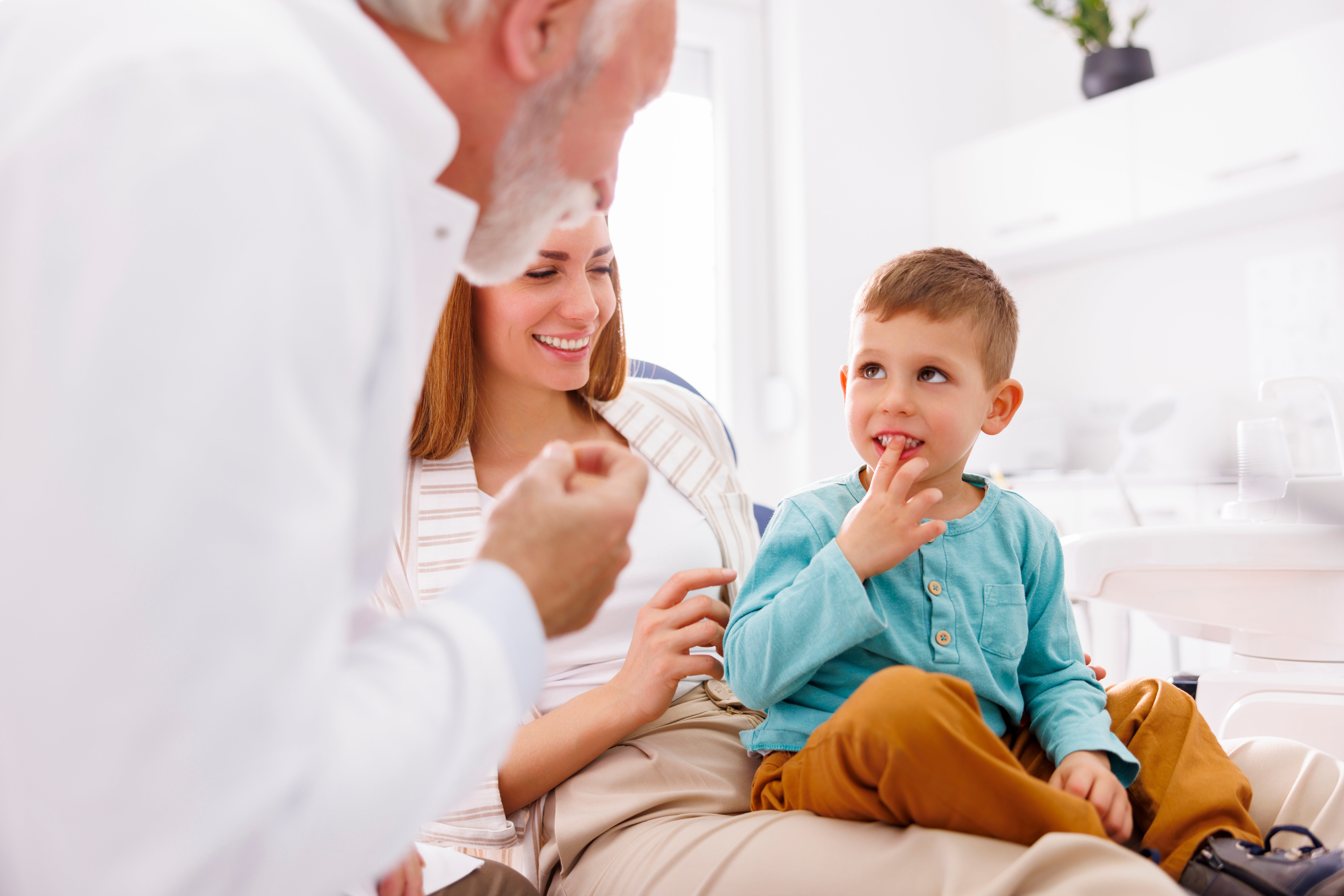 Young boy sitting on a woman's lap in a dental office, showing his teeth to an older male dentist.