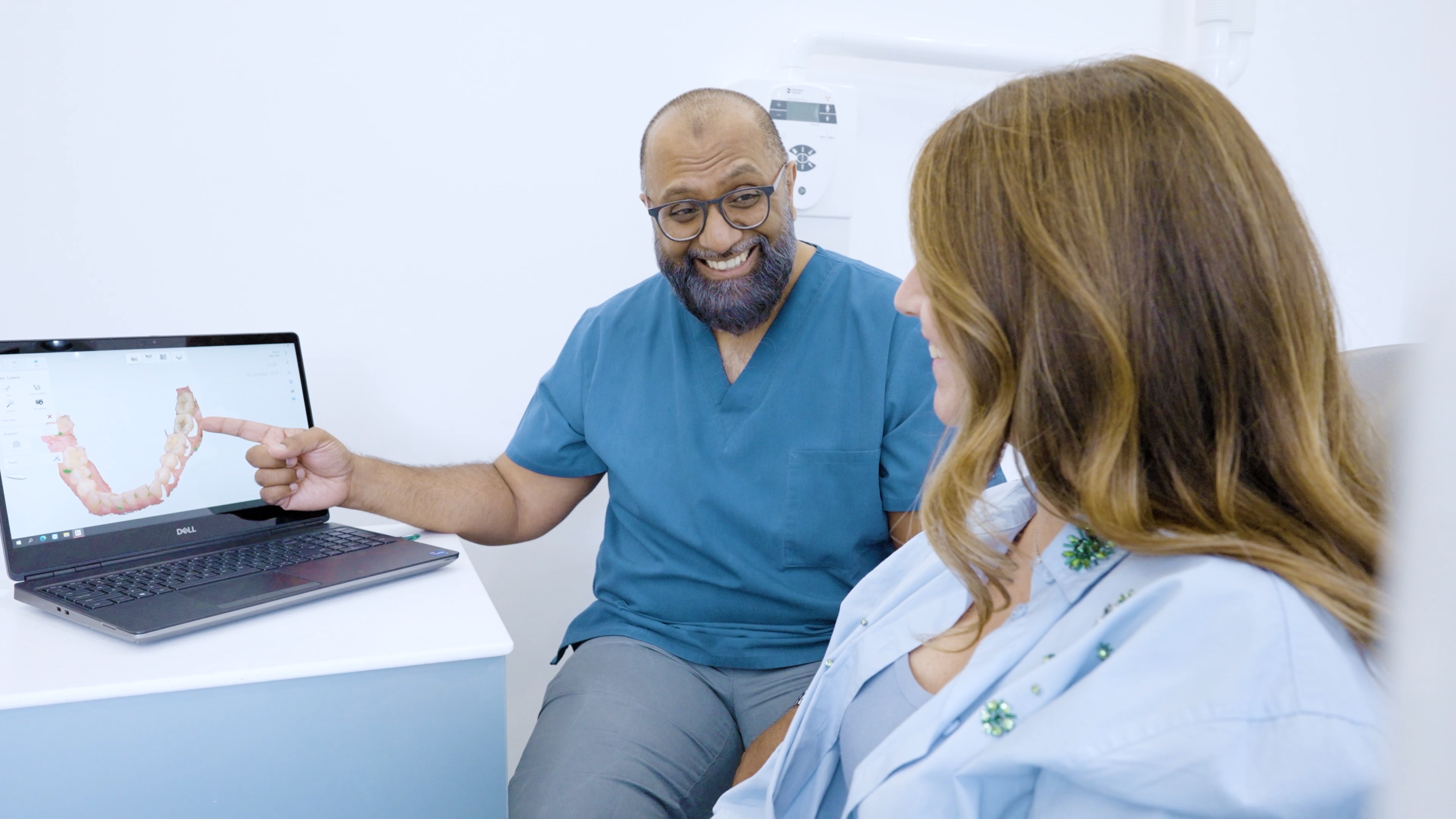 Dentist in blue scrubs smiling and pointing at dental scan on laptop screen with female patient.