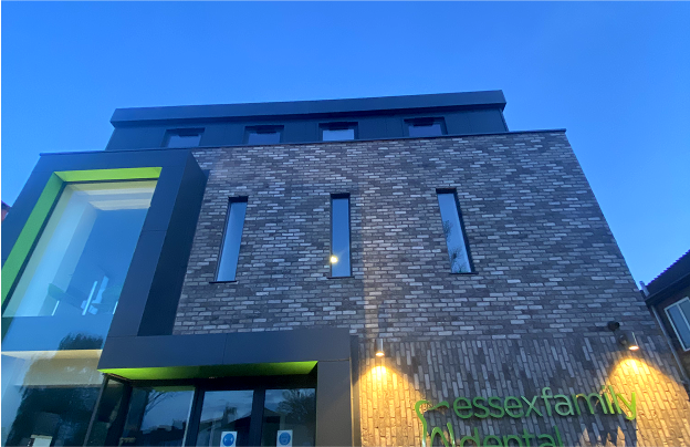 Modern brick building with narrow vertical windows and illuminated green signage that reads 'Essex Family Dental' during dusk.