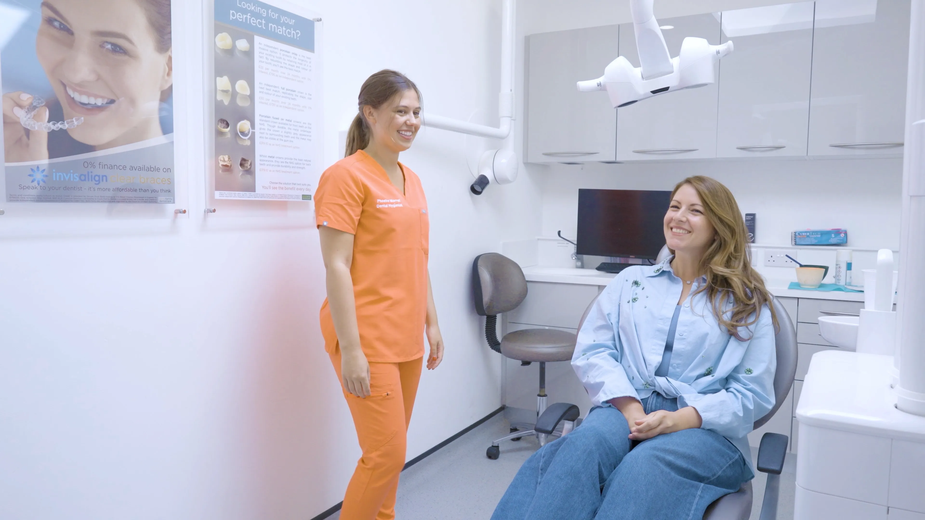 Smiling female dentist in orange scrubs stands next to a happy woman seated in a dental chair in a bright clinic room.