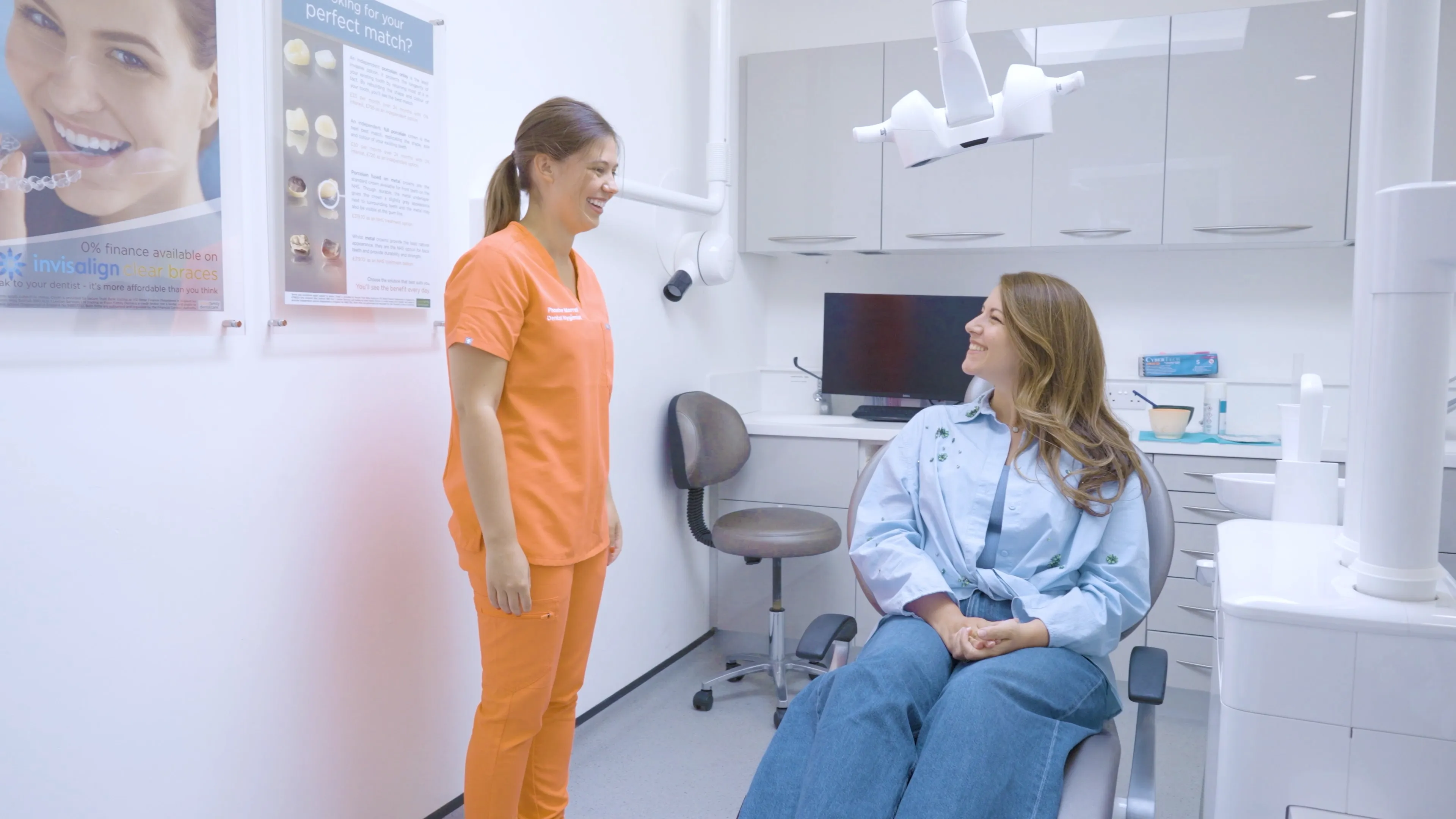 Dentist in orange scrubs smiling and talking to a patient seated in a dental chair in a modern dental office.