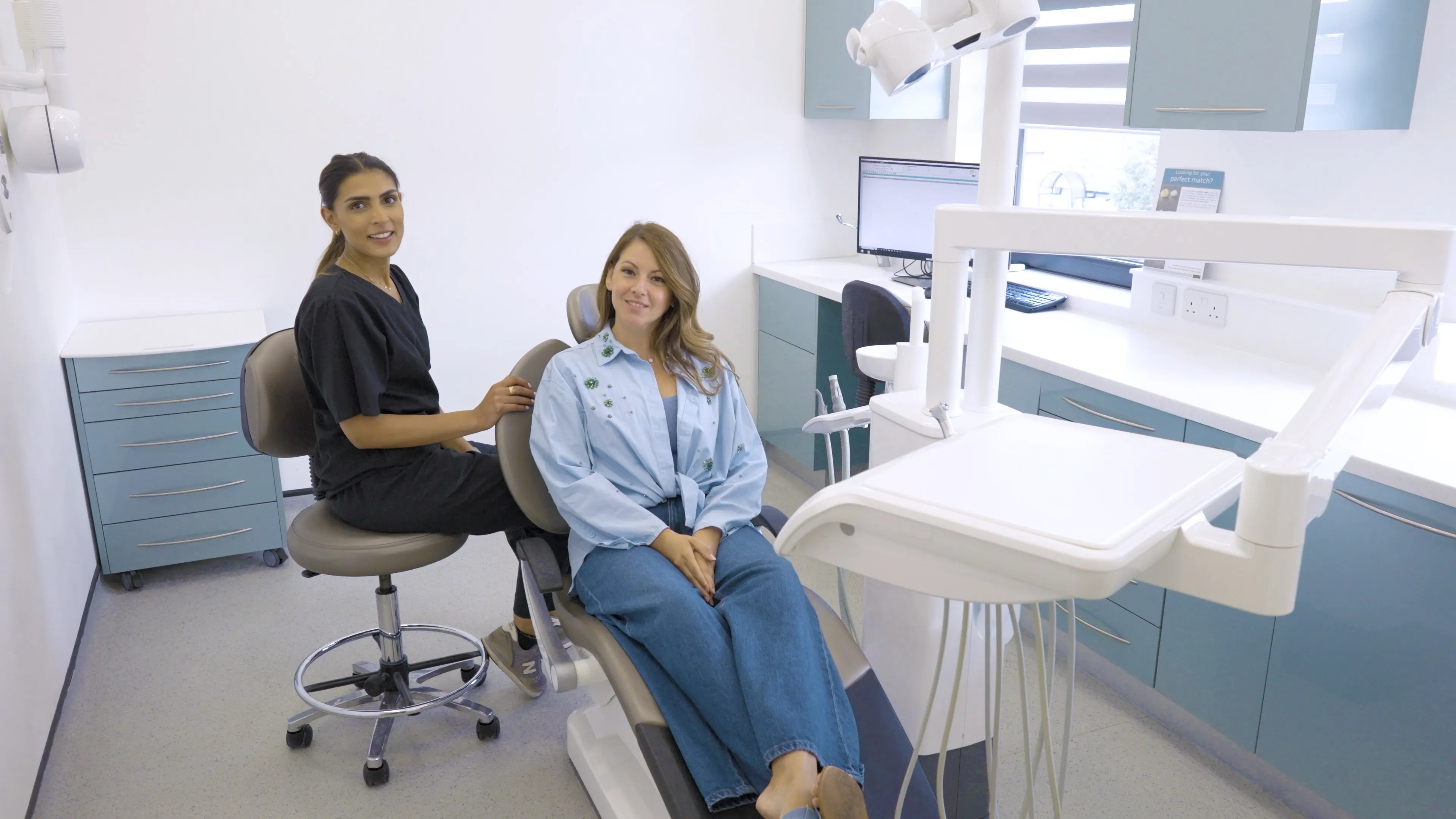 Dentist in black scrubs sitting on a stool next to a dental patient seated in a dental chair in a modern clinic room.