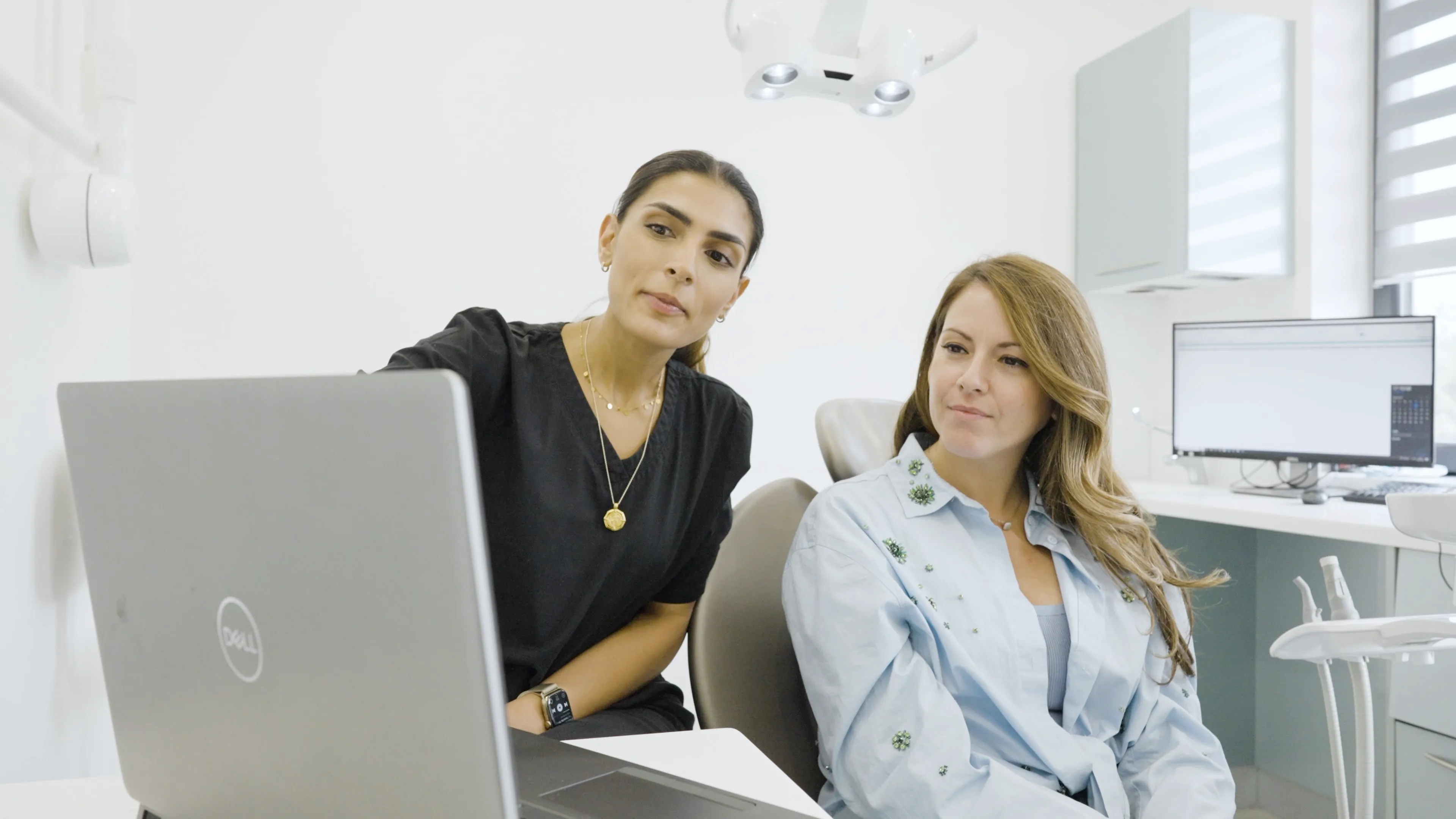 A medical professional in black scrubs explaining information on a laptop to a patient seated in a dental chair.