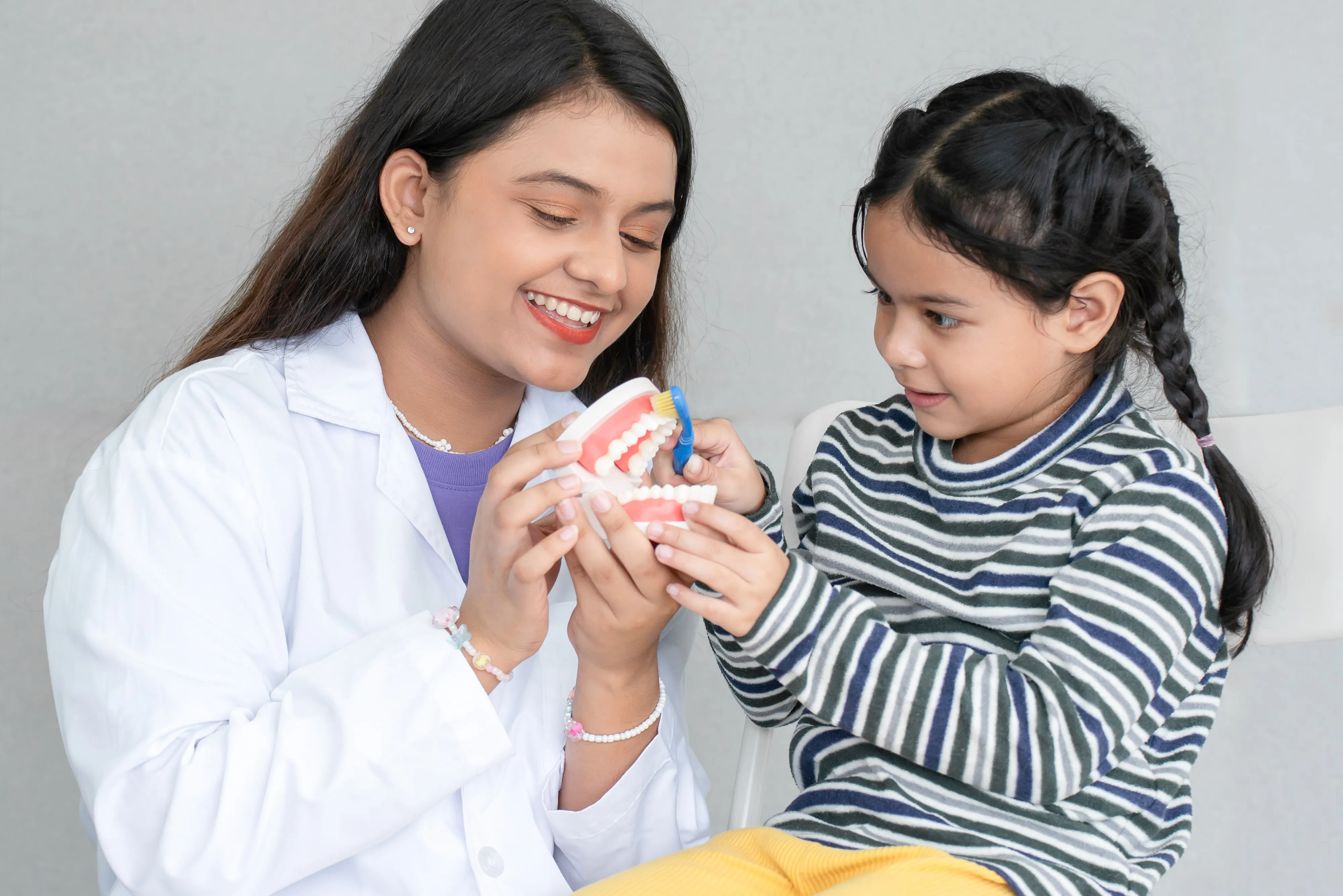 Smiling female dentist teaching a young girl how to brush teeth using a dental model and toothbrush.