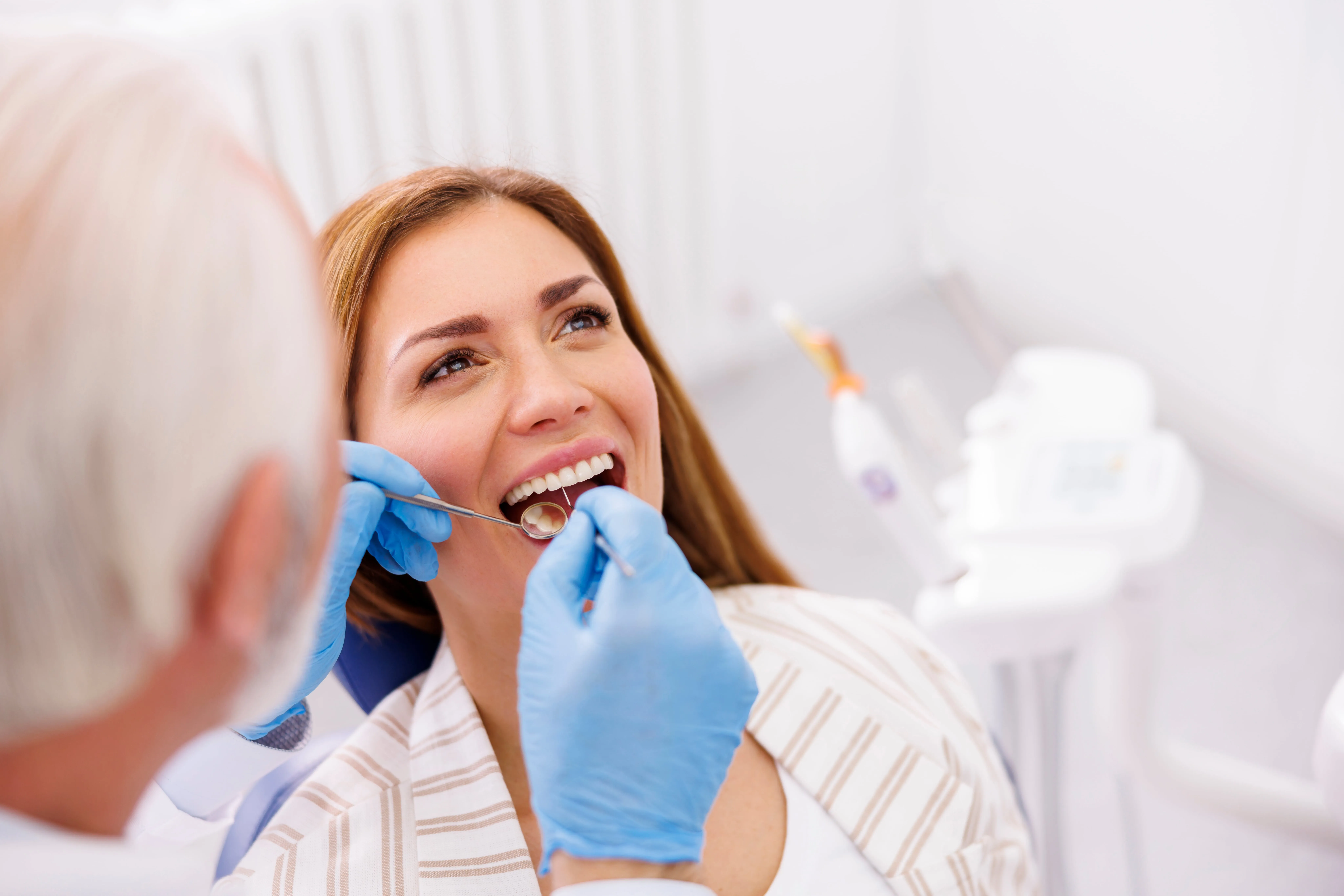 Dentist wearing blue gloves examining a smiling woman's teeth with dental tools in a bright clinic.