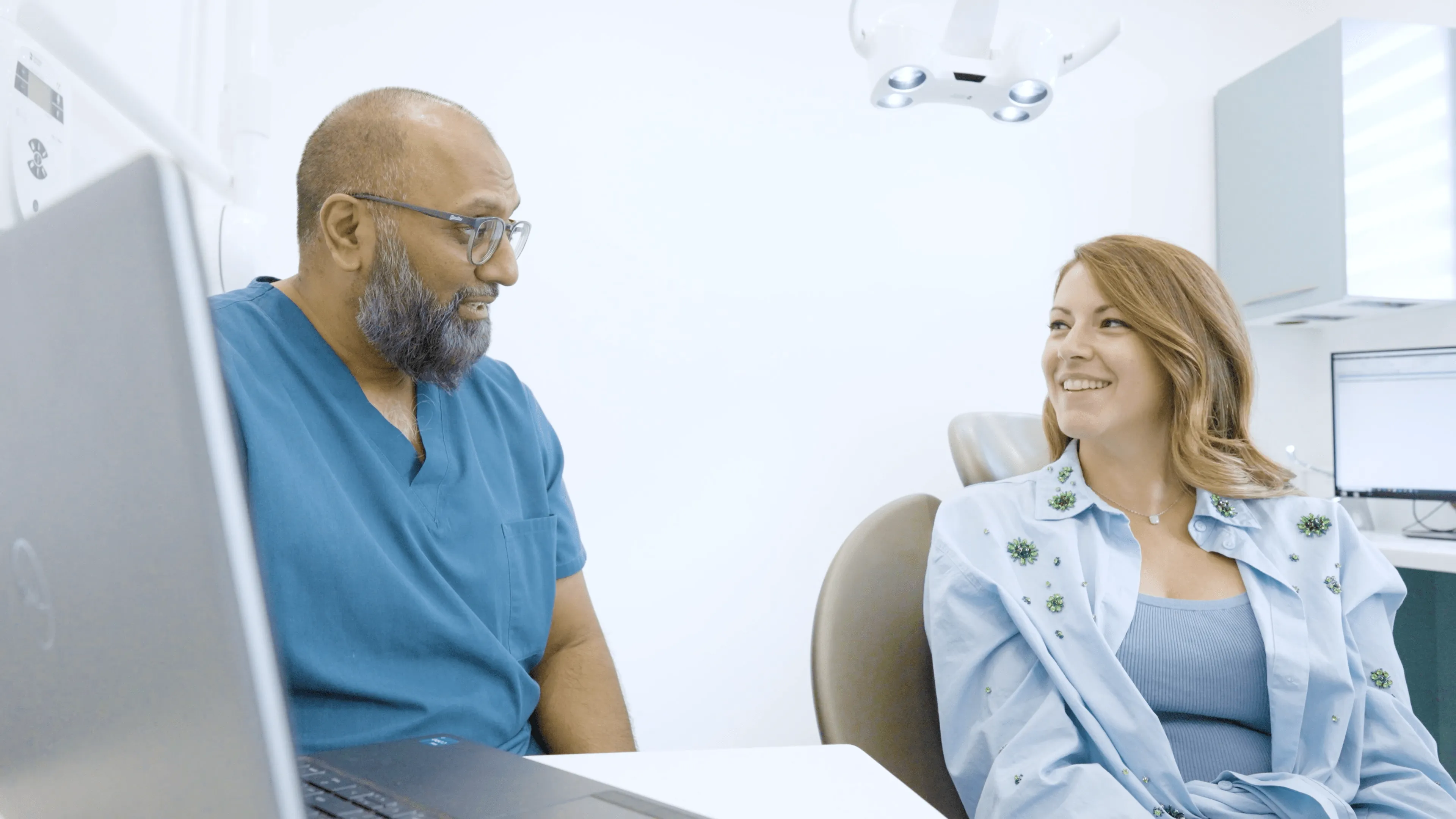 Male doctor in blue scrubs talking to a smiling female patient seated in a medical office.
