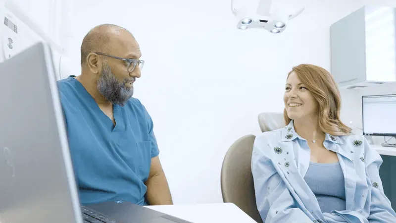 Dentist in blue scrubs smiling and showing a dental scan on a laptop to a female patient in a light blue shirt.