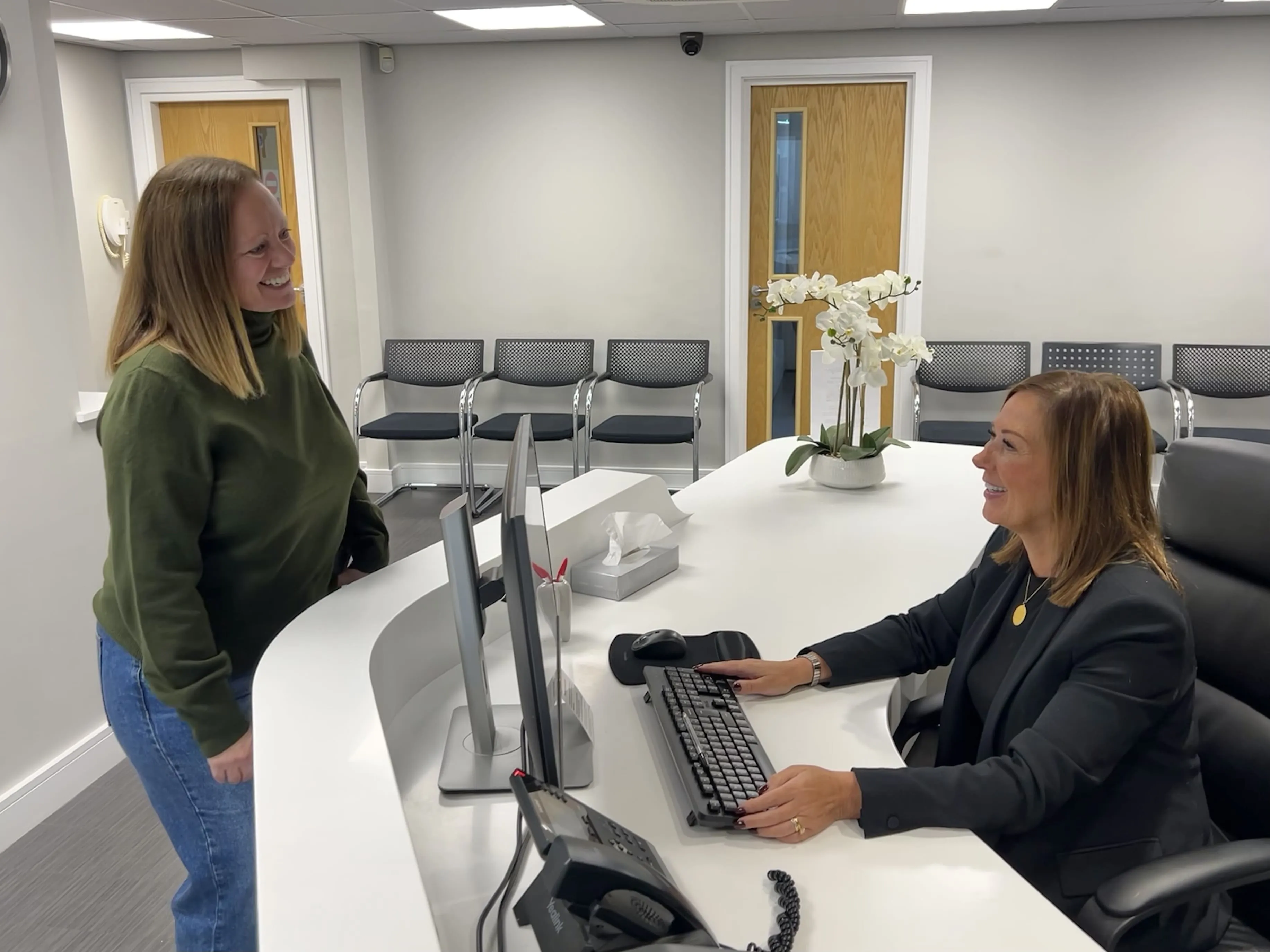 Receptionist sitting at a desk with two computer monitors smiling and talking to a standing visitor in a green sweater and blue jeans in a waiting area.