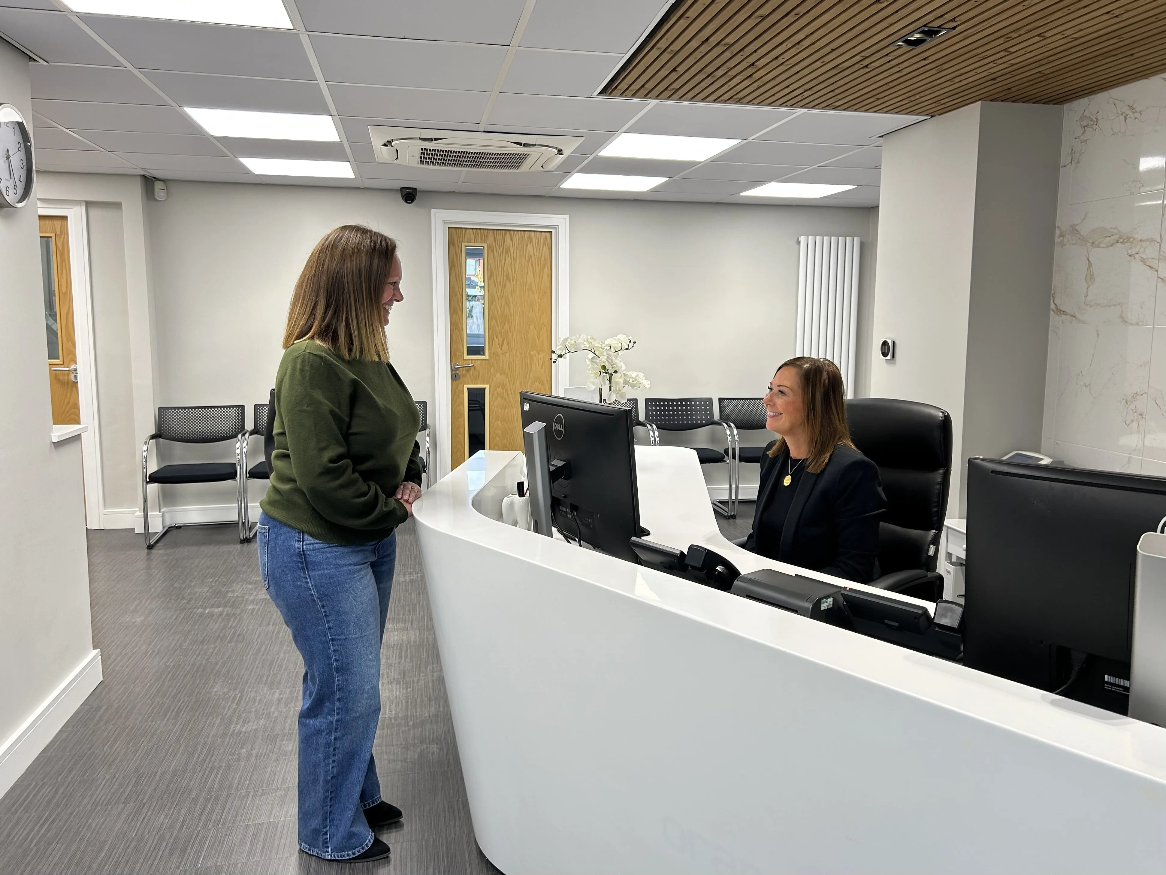 A woman in a green sweater standing at a white reception desk talking to a receptionist seated at a computer in a modern office lobby.