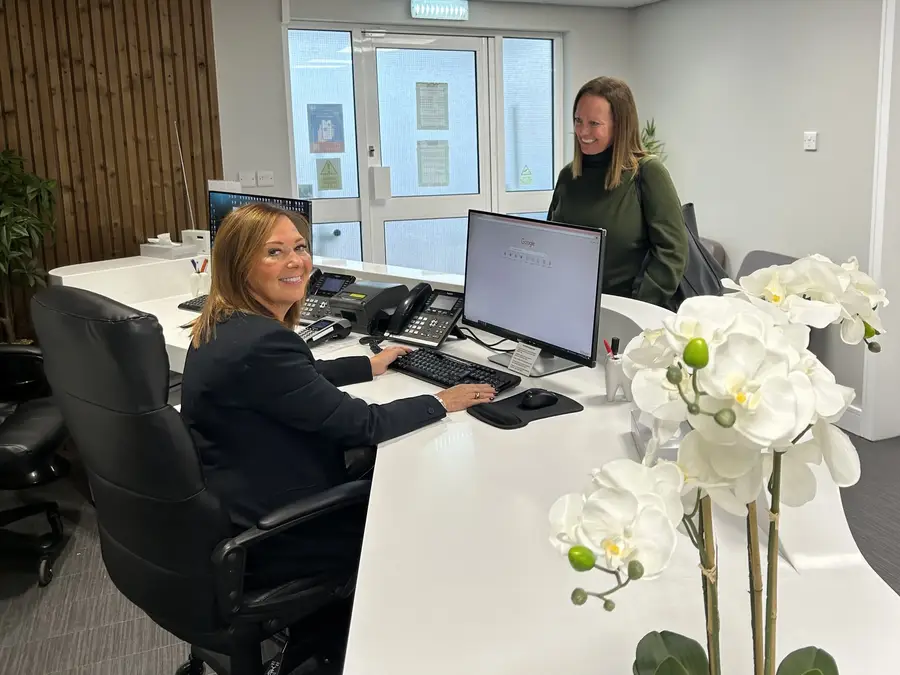 Receptionist sitting at desk smiling at a standing visitor near white orchids in a modern office.