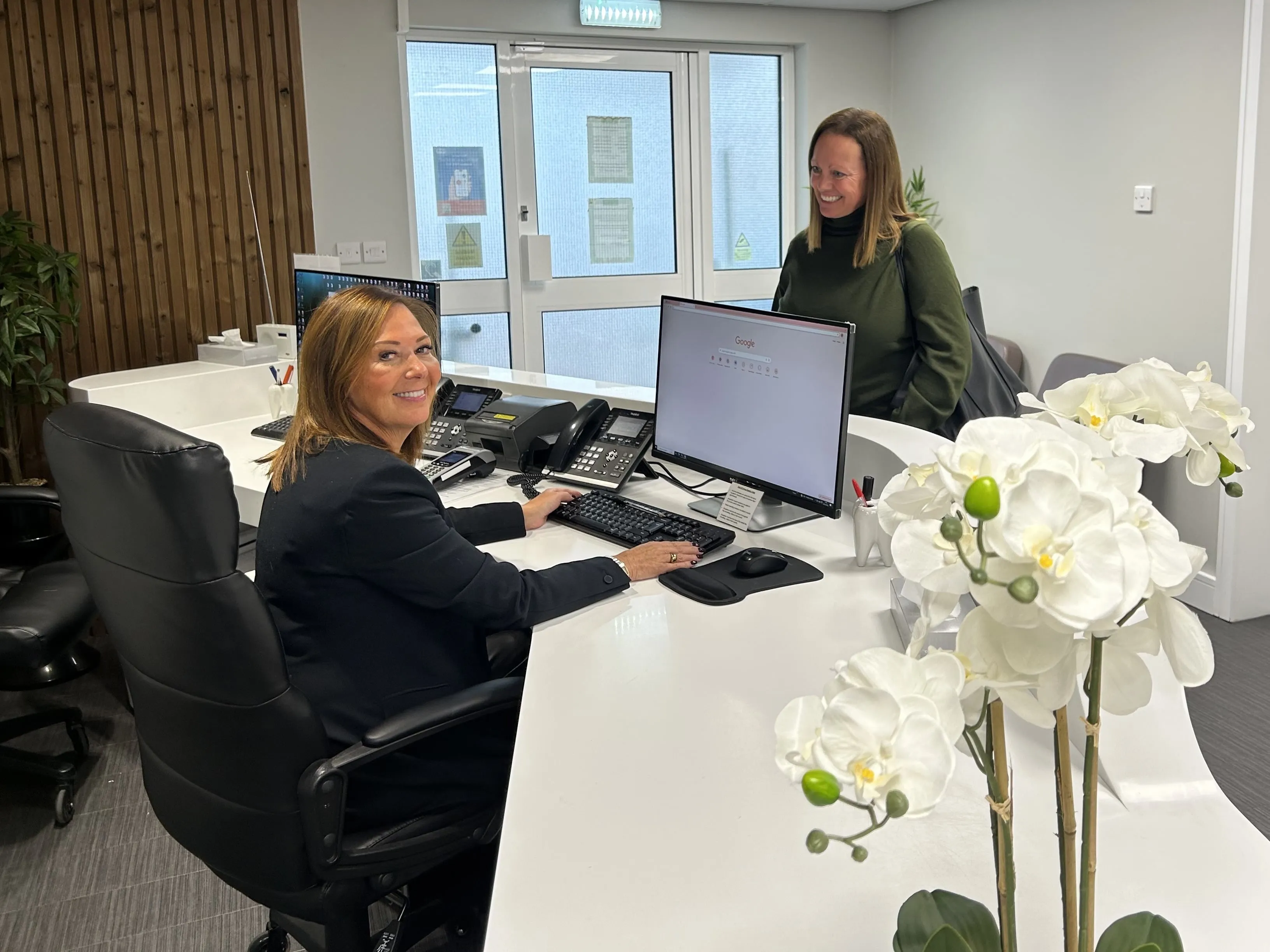 Receptionist sitting at desk smiling at a standing visitor near white orchids in a modern office.