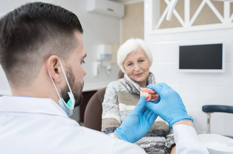 Dentist wearing blue gloves demonstrating dentures to an elderly woman in a dental clinic.