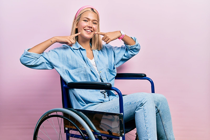 Smiling young woman sitting in a wheelchair points to her cheeks against a pink background.