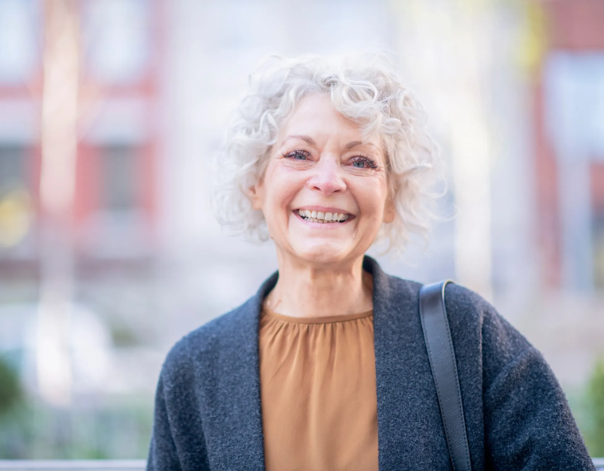 Smiling elderly woman with curly white hair wearing a brown top and dark cardigan outdoors.