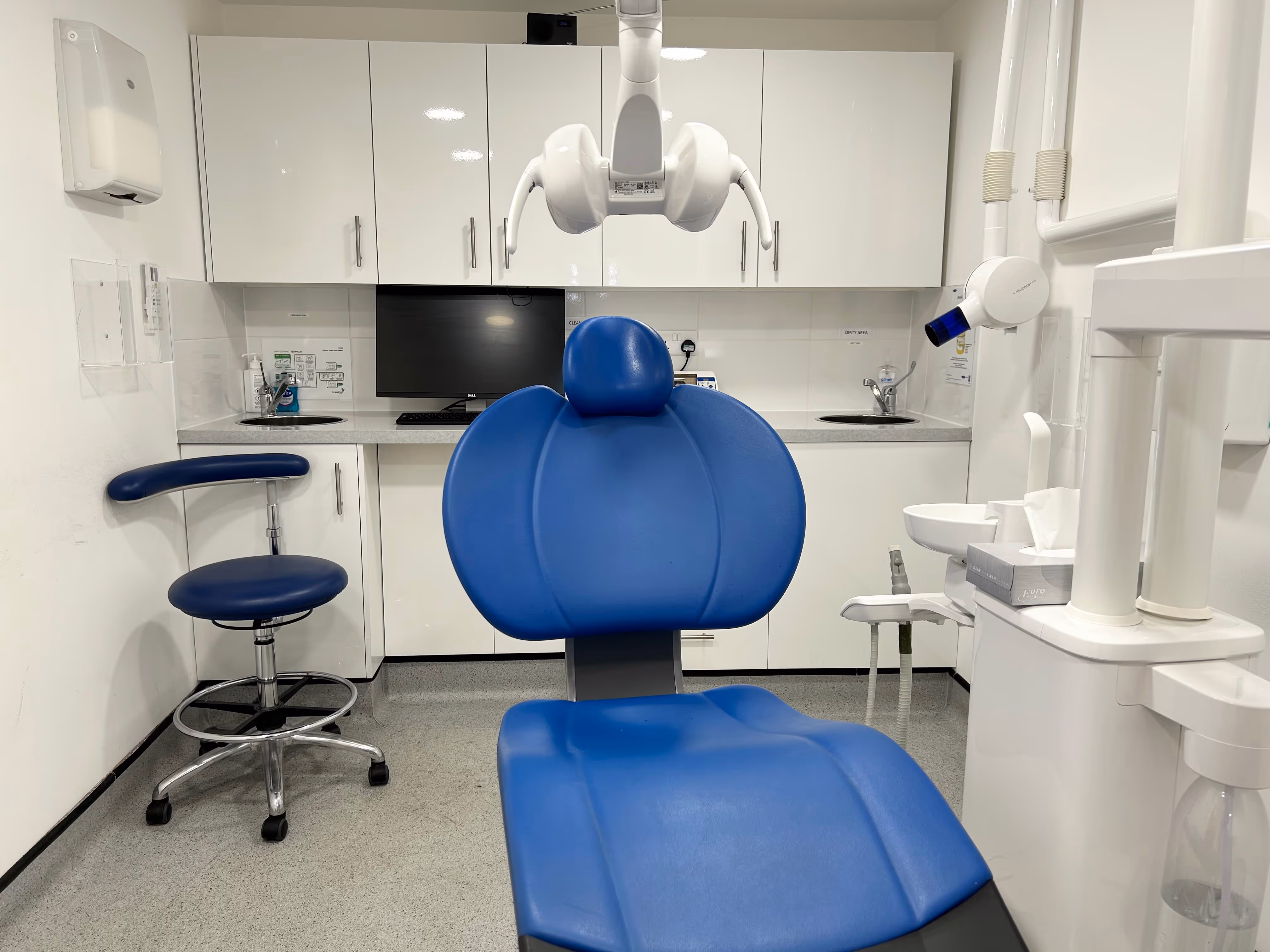 Modern dental clinic room with a blue dental chair, a matching stool, white cabinetry, a computer screen, and dental equipment.