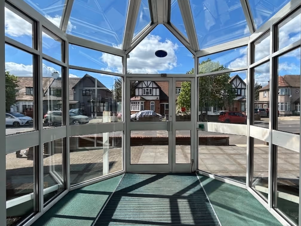 View from inside a glass-walled, polygonal entrance vestibule looking out onto a street with parked cars and suburban houses under a blue sky with scattered clouds.