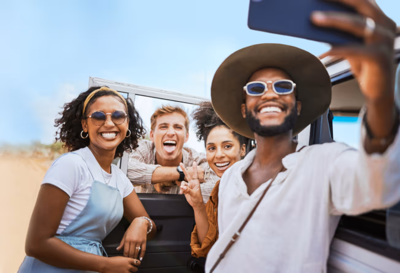 Four friends smiling and taking a selfie beside a car on a sunny day.
