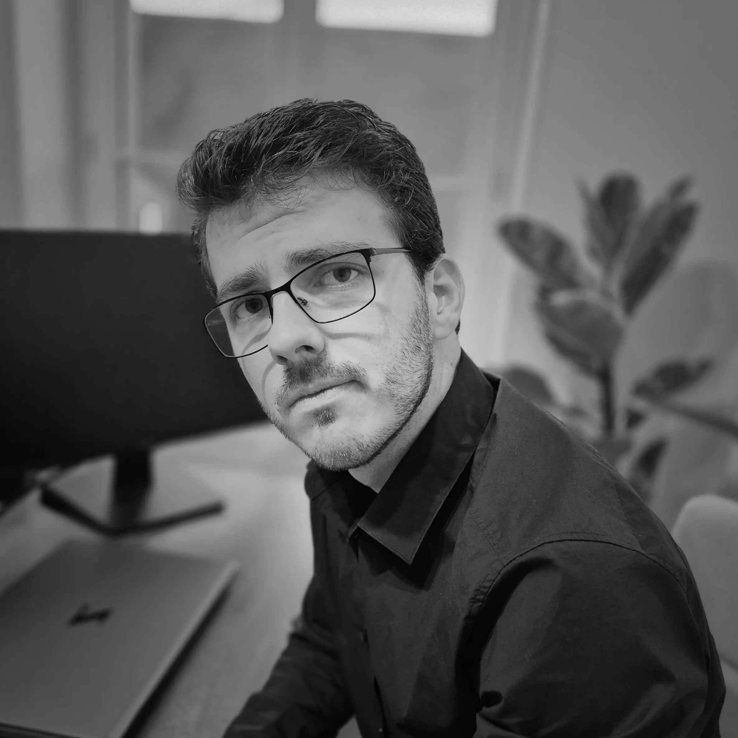 Homme portant des lunettes en chemise noire assis devant un ordinateur portable sur un bureau avec une plante floue en arrière-plan.