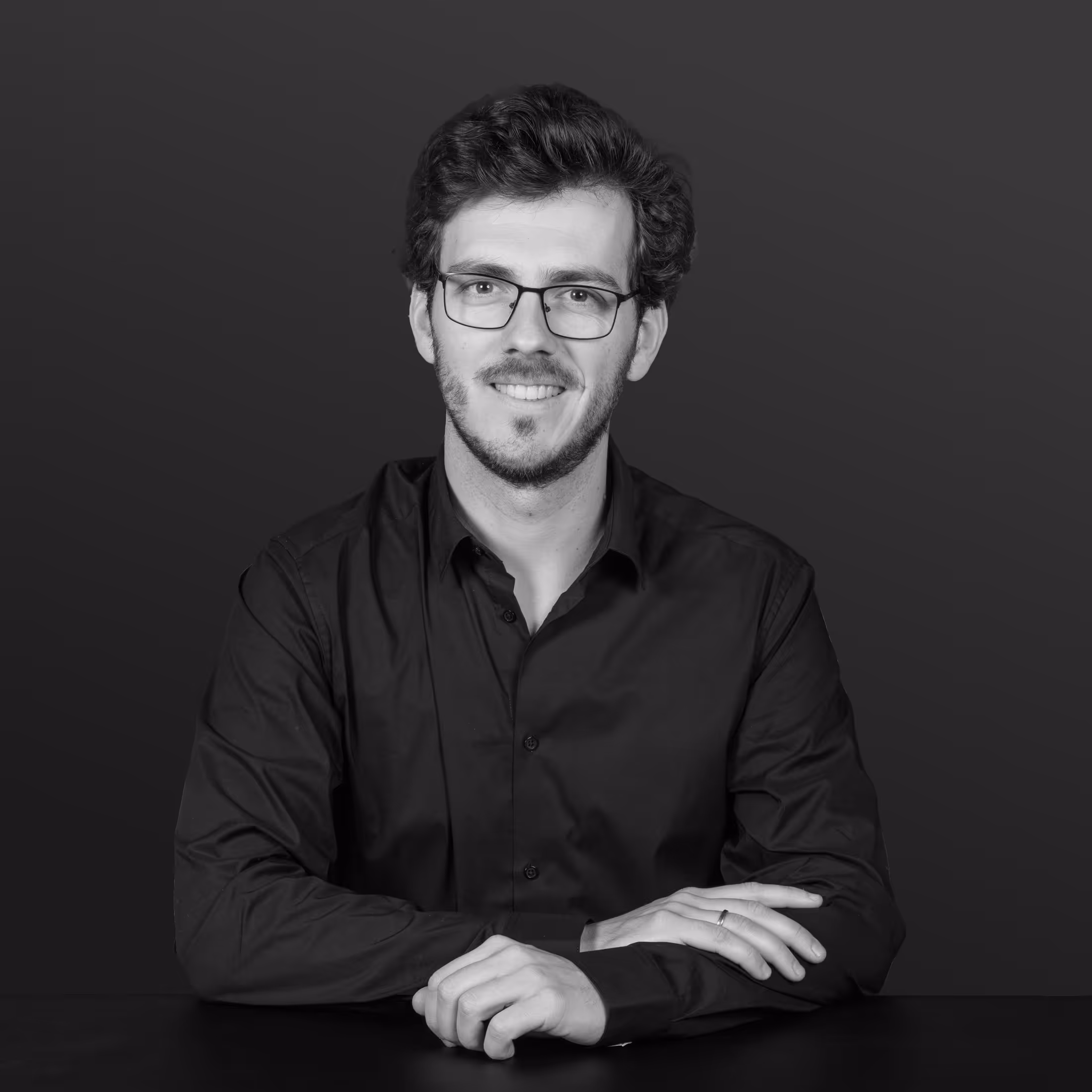 Homme portant des lunettes en chemise noire assis devant un ordinateur portable sur un bureau avec une plante floue en arrière-plan.